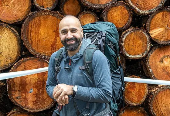 A man from a Pakistani family background on a outdoor walk, wearing hiking gear. He stands in front of a pile of logs