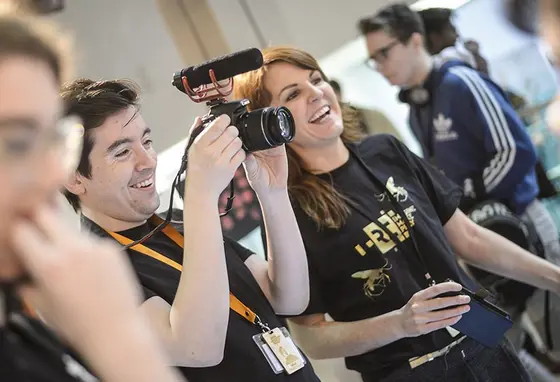 Two people wearing Reimagine, Remake, Replay tshirts using filming equipment at an event