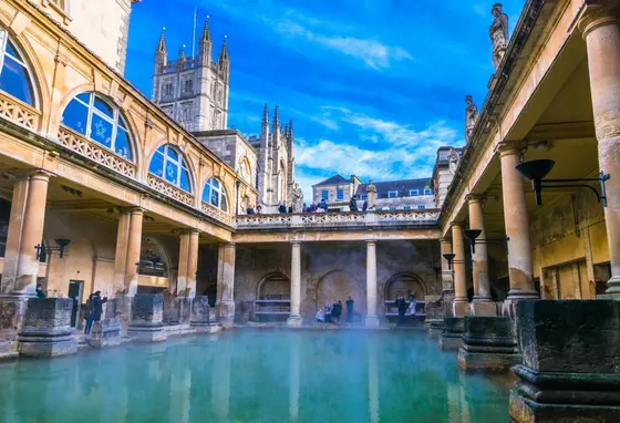 Looking across the water at the Roman Baths in Bath, people standing on the edges and looking down from a walkway above with 
