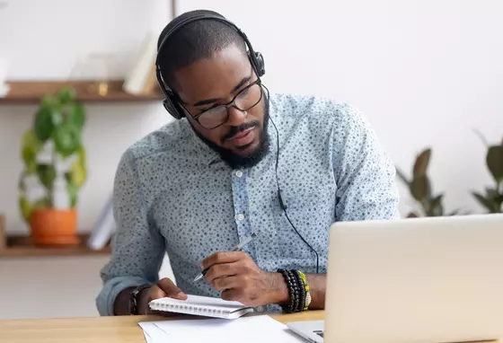 A black man waering headphones, looking at a laptop computer and taking notes in a notebook