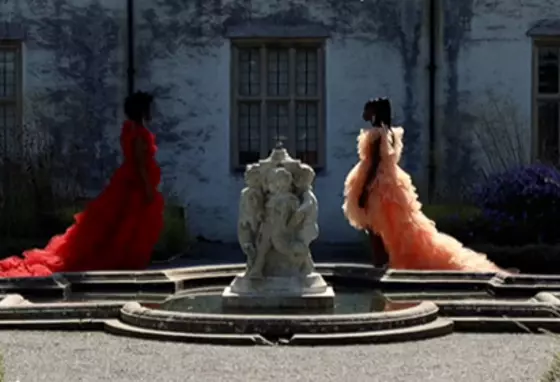 Two people dressed in colourful dresses walk towards a fountain outside a historic house