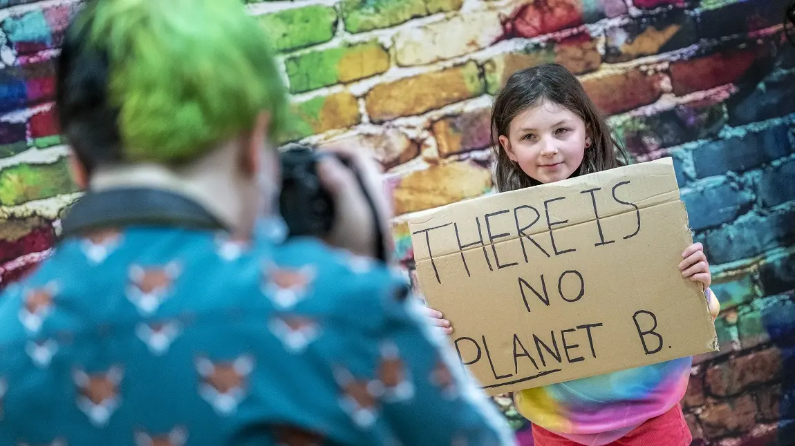 A child having their photograph taken, holding up a cardboard sign that reads: There is no planet B