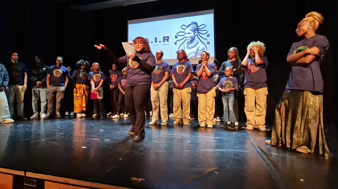A large group on stage with a person reading from a sheet of paper in the foreground