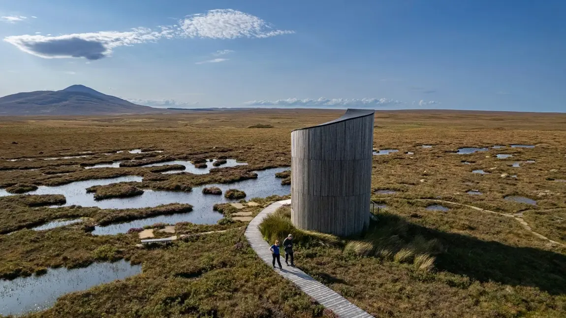 A remote, contemporary bird-watching tower in the middle of vast wetlands. Across a wooden causeway, two birdwatchers depart the tower.