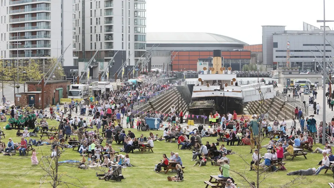 Hundreds of people are gathered, enjoying the sunshine, around a historic ship in dry dock on the Belfast quayside.