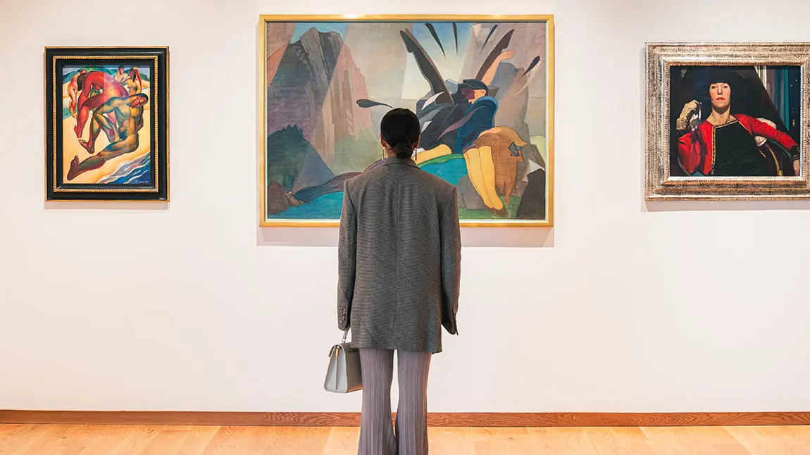A young person looking at one of three paintings on a gallery wall. The person has their back to us.