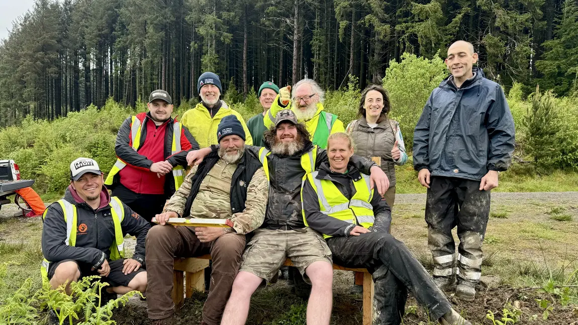 A group of men and women in high vis jackets and outdoor attire gather around a newly built bench on a wooded hillside.