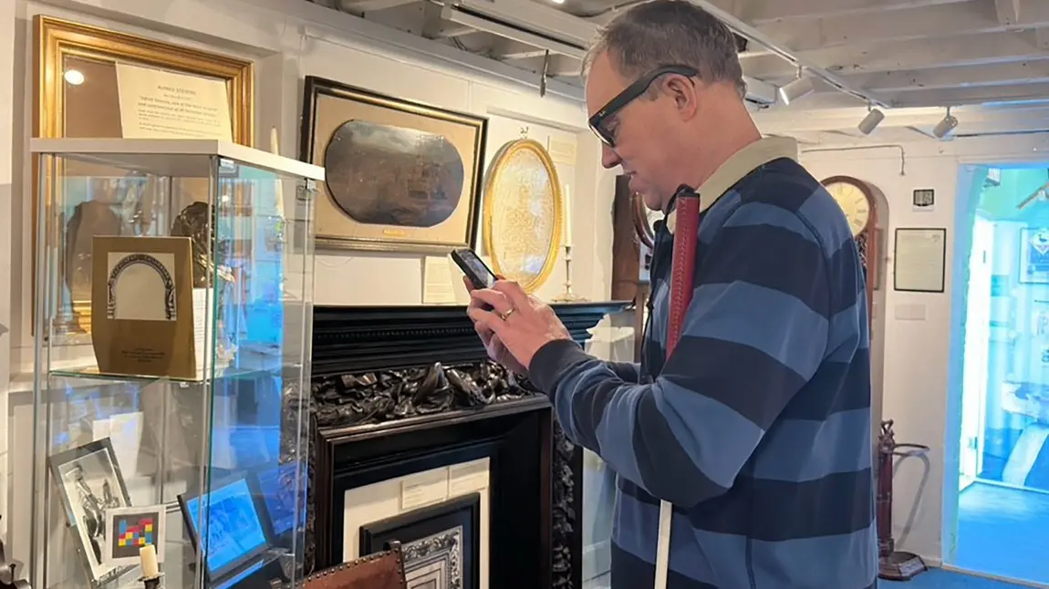 A person holding a white cane and using a smartphone, in front of them is a glass museum case containing old photographs and a brightly coloured NaviLens code