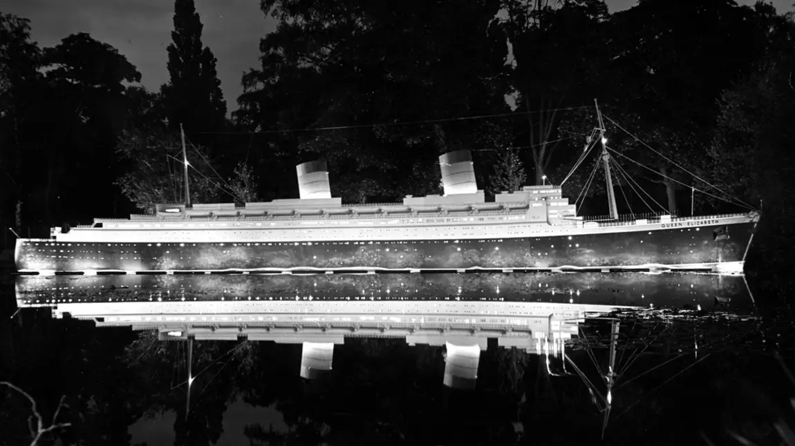 An illuminated replica of the ship Queen Elizabeth floats on the park lake at night.