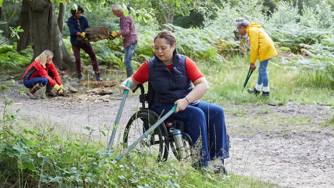 A volunteer in a wheelchair using shears to do practical work in woodland. Other volunteers can be seen doing other practical tasks in the background
