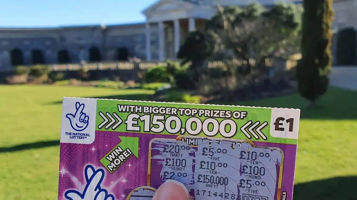 A National Lottery scratchcard held in front of a large building with classical architecture and lawn 
