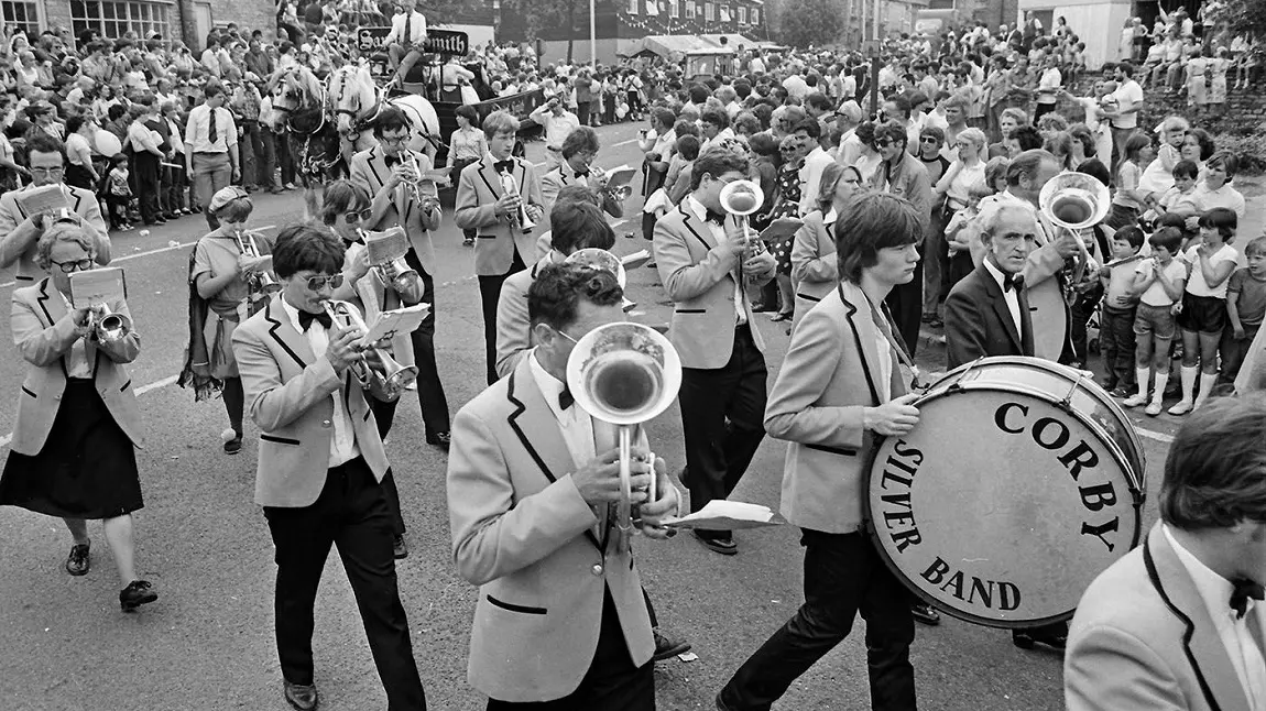 A black and white image of a brass band marching through the streets of a packed town centre.