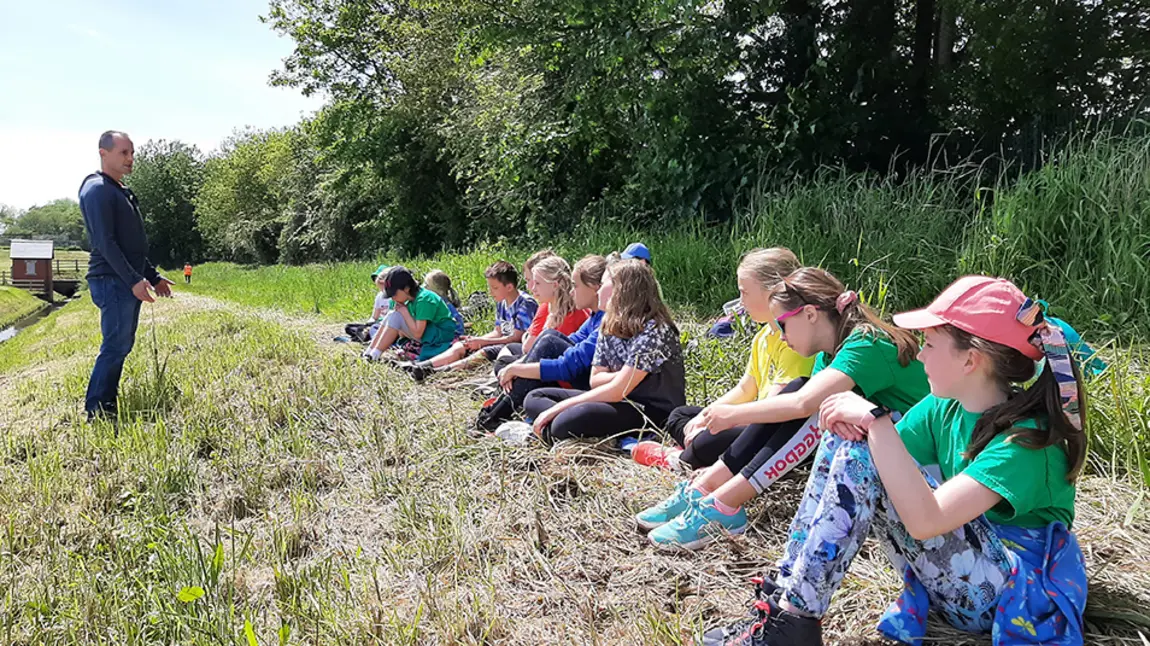 A standing man talking to children sitting on the edge of woodland