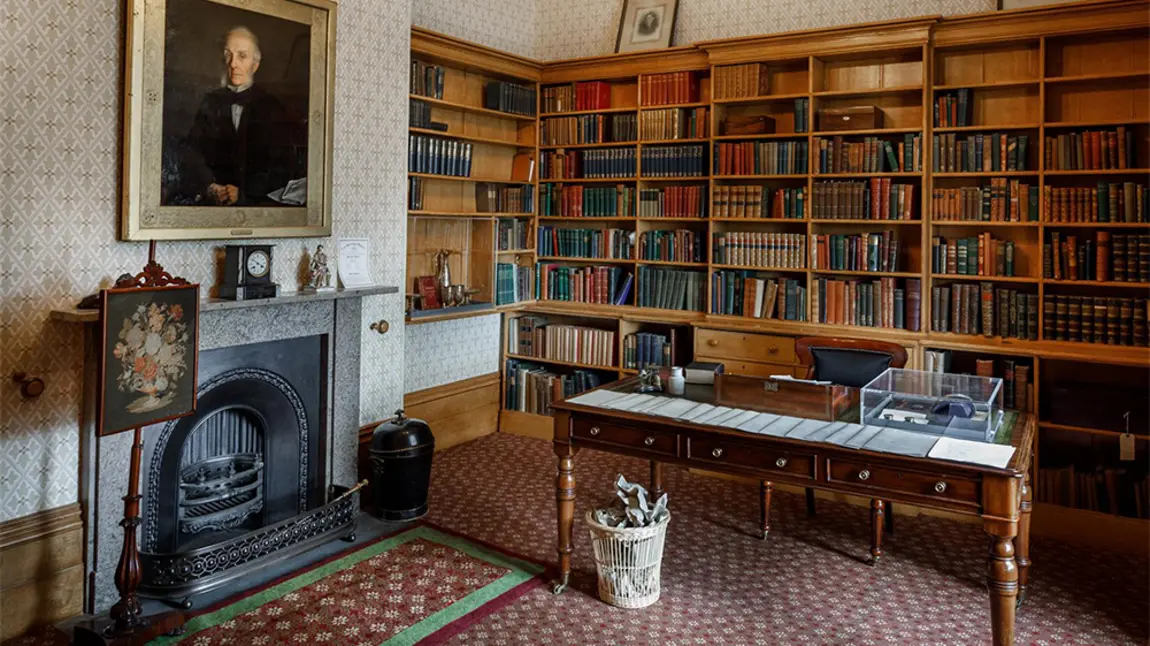 Inside a room with desk, book case and fire place in background