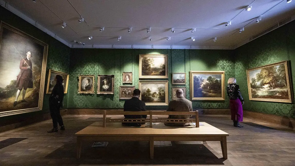 A gallery space with people observing paintings and sitting down