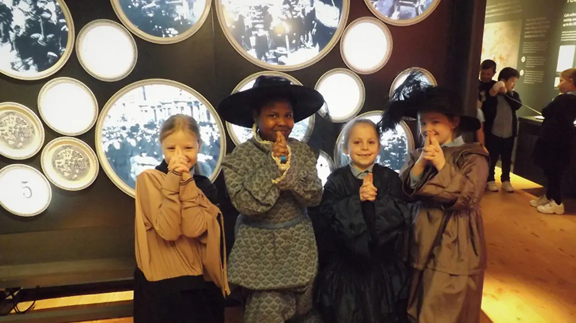 Four children face camera smiling. They are dressed in historic outfits. They are near an exhibition. 