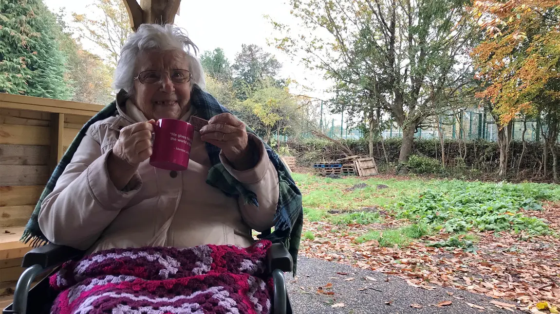 Elderly woman using wheelchair in garden, smiling with tea and biscuit