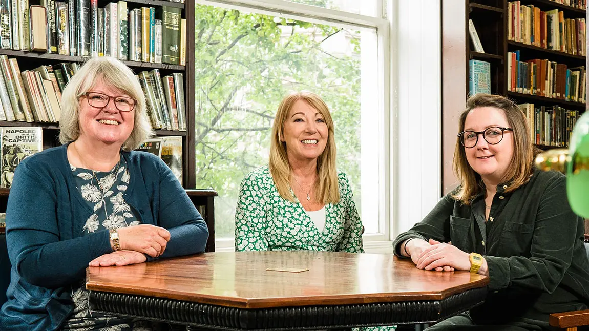 Three people sit at table in room surrounded by books on shelves. They smile at the camera.