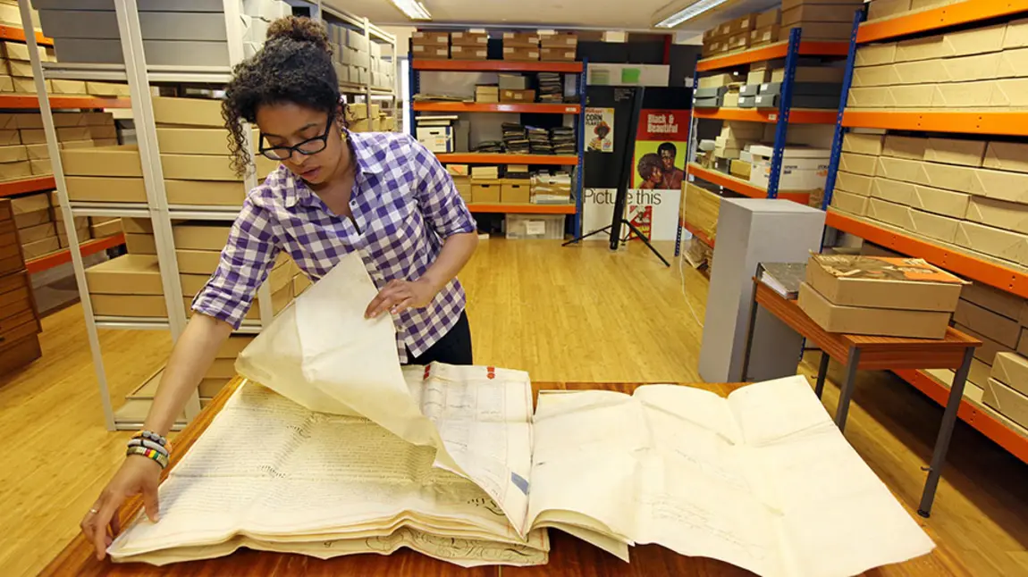 A person handles large parchment documents in an archive room, with shelves of archive boxes in the background