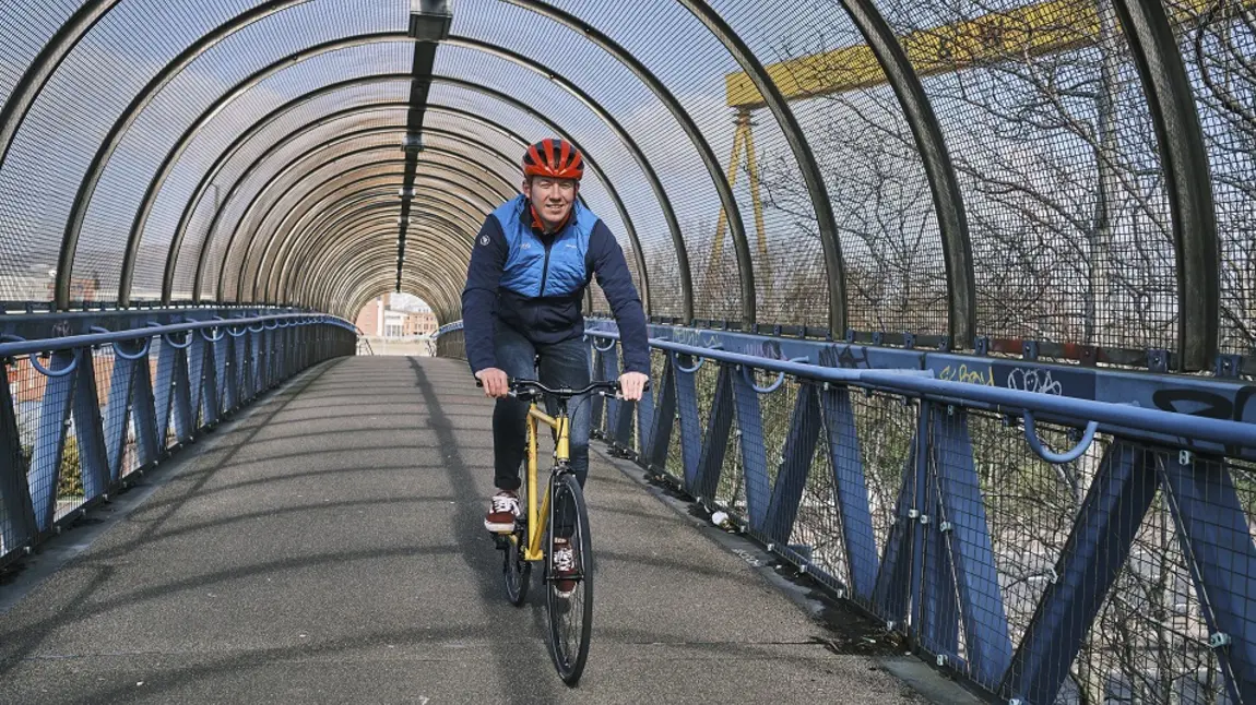A man cycling on a bridge with the Harland and Wolff cranes visible in the background