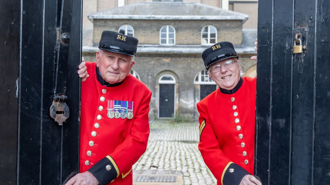 Two people wearing scarlet Chelsea Pensioners' uniform open stable doors at Royal Chelsea Hospital, London with historic building and courtyard behind