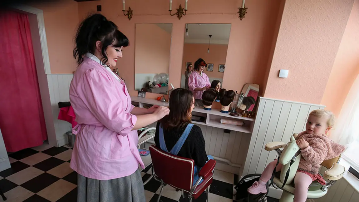 The interior of a 1950s hairdresser's shop. A visitor is having their hair styled by a stylist in vintage clothing. A child sits in a high chair next to the visitor.