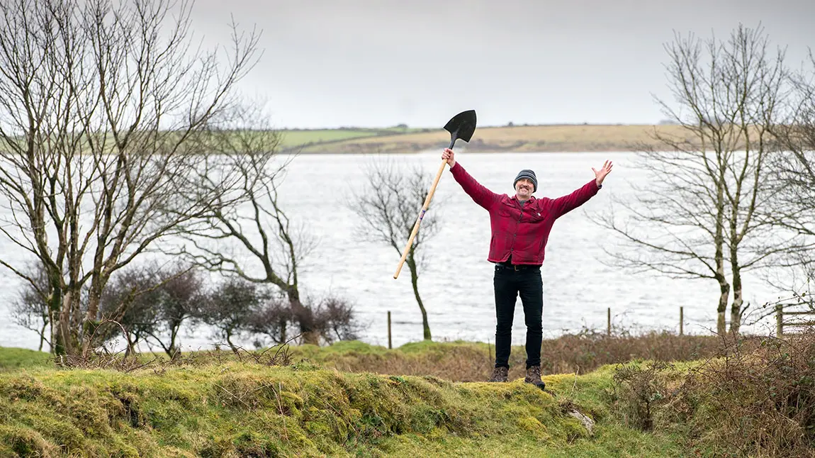 Person yn sefyll ym myd natur yn edrych yn hapus wrth ddal rhaw yn yr awyr