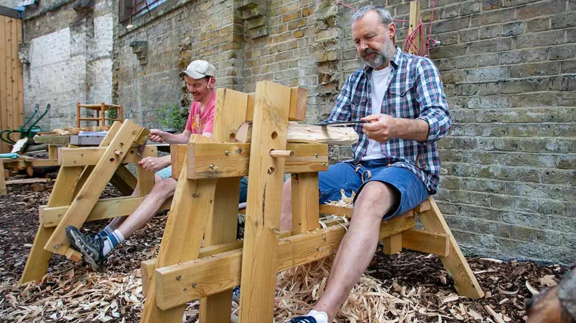 Two people sitting outside carving wood at woodworking benches