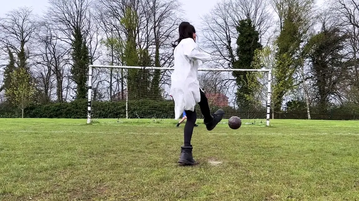 A young girl in a white dress and boots takes a penalty kick.