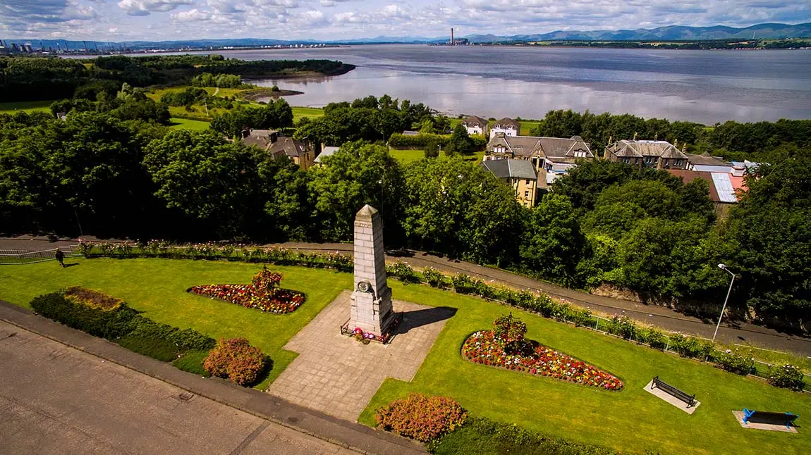 Bo'ness war memorial is at the centre of the frame and behind it there are trees and houses. In the distance is the Firth of Forth.