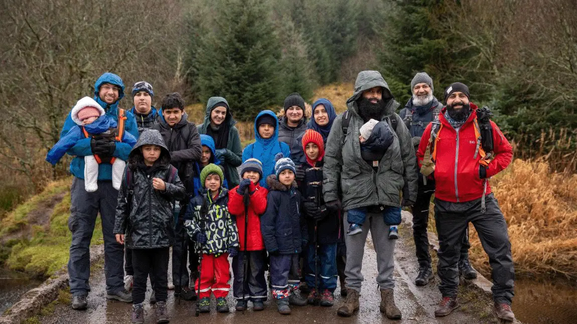 A large group of people of all ages in walking gear on a rainy day. They are standing on a path through a wooded glen.