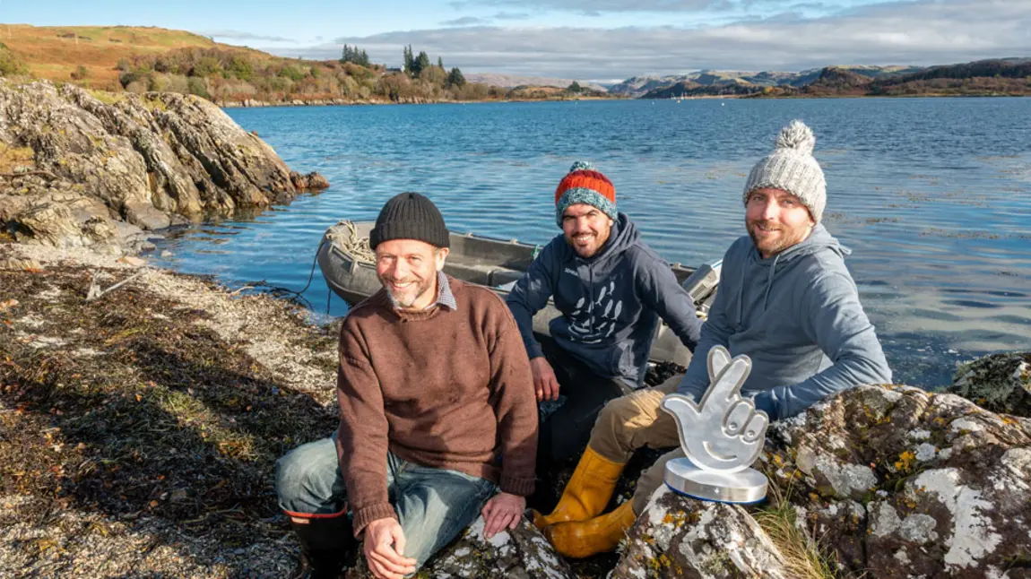 Three people sit on the edge of a loch in Scotland, with a boat in the background and the award in front of them