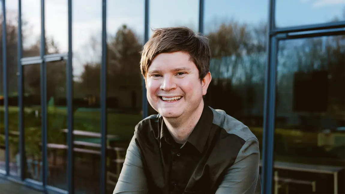 a photo of David Sheppeard sitting outside against a glass wall. David has white skin, short brown hair and wears a dark shirt