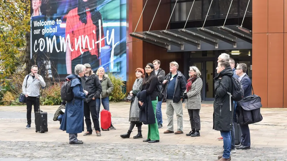A group of 13 people standing outside a building listening to a tour guide as part of a community walking tour
