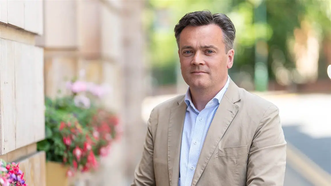 James Twining wearing shirt and jacket, standing next to a stone building with flower boxes.