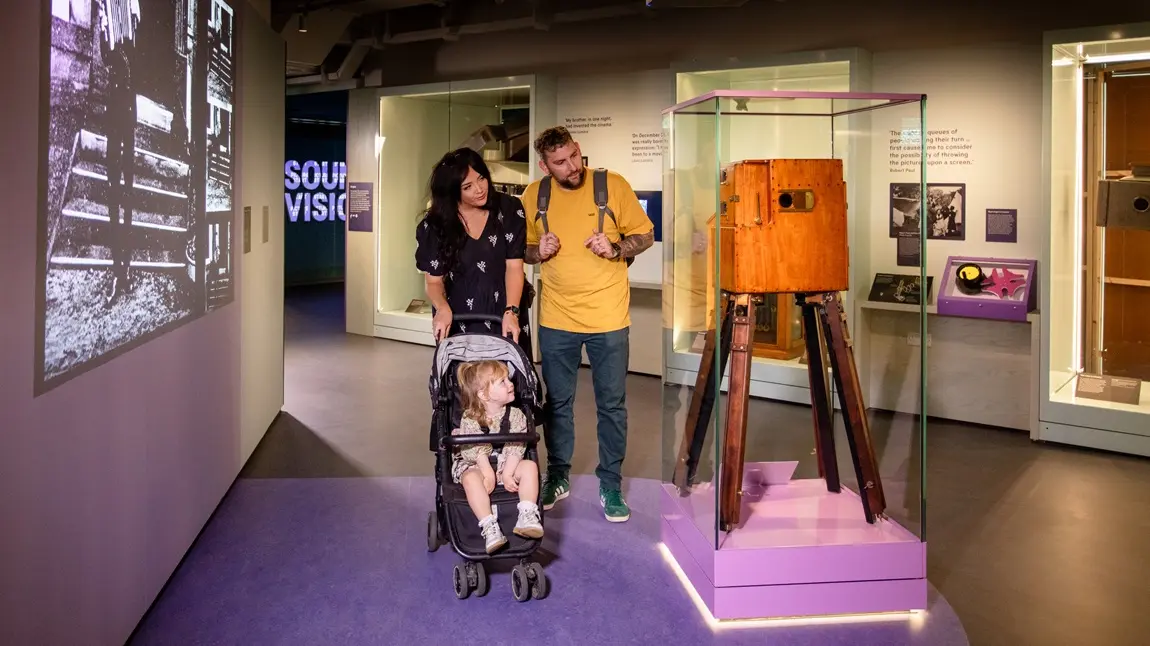 A man and a woman push a child in a buggy as they take a closer look at a pioneering film camera from the early days of the technology. A black and white film of the era is projected on the wall to their left.