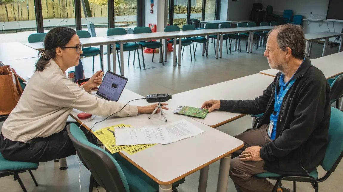 Two people sit at a desk with a laptop and microphone to record the interview