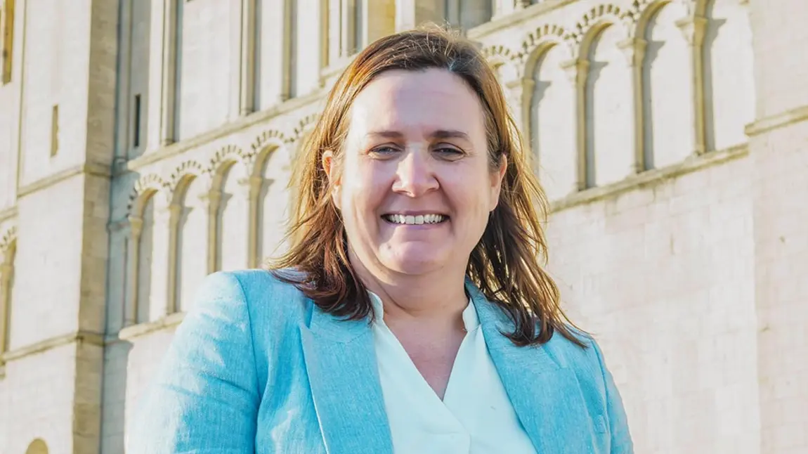 A head and shoulders photo of Liz Bates, a woman with fair skin and brown hair wearing a suit, standing in front of Norwich Castle
