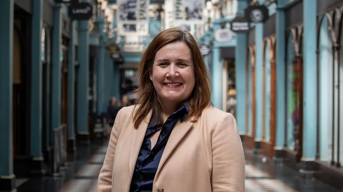 Liz Bates A photo of Liz Bates, a woman with fair skin and brown hair wearing a blue silk shirt and cream coat, standing in a historic shopping arcade