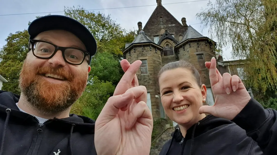 A man and a woman are stood in front of a heritage site doing the crossed fingers selfie