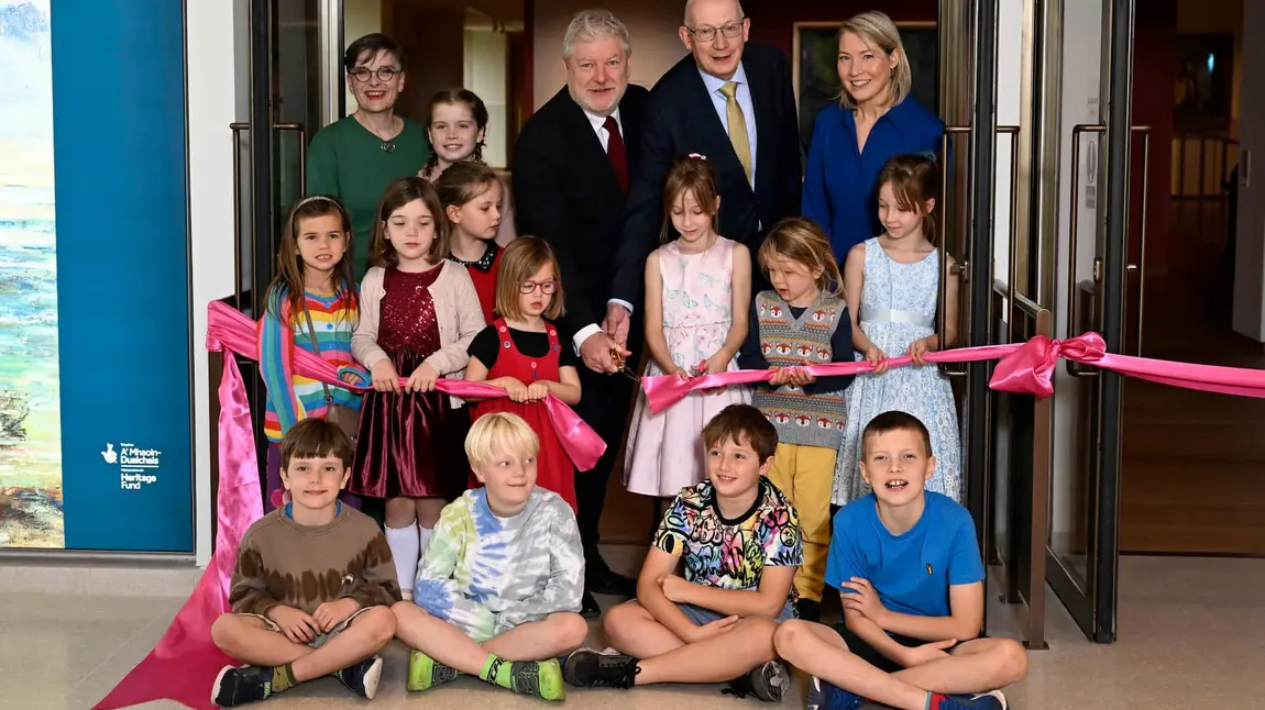 A group of children and adults sit and stand as they pose for a photo while cutting a ribbon.