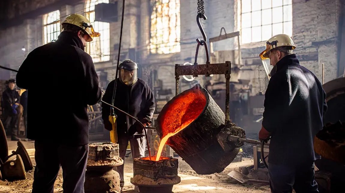 Three people stand inside a factory casting a large bell. Molten metal pours from a container held in the air by a hoist