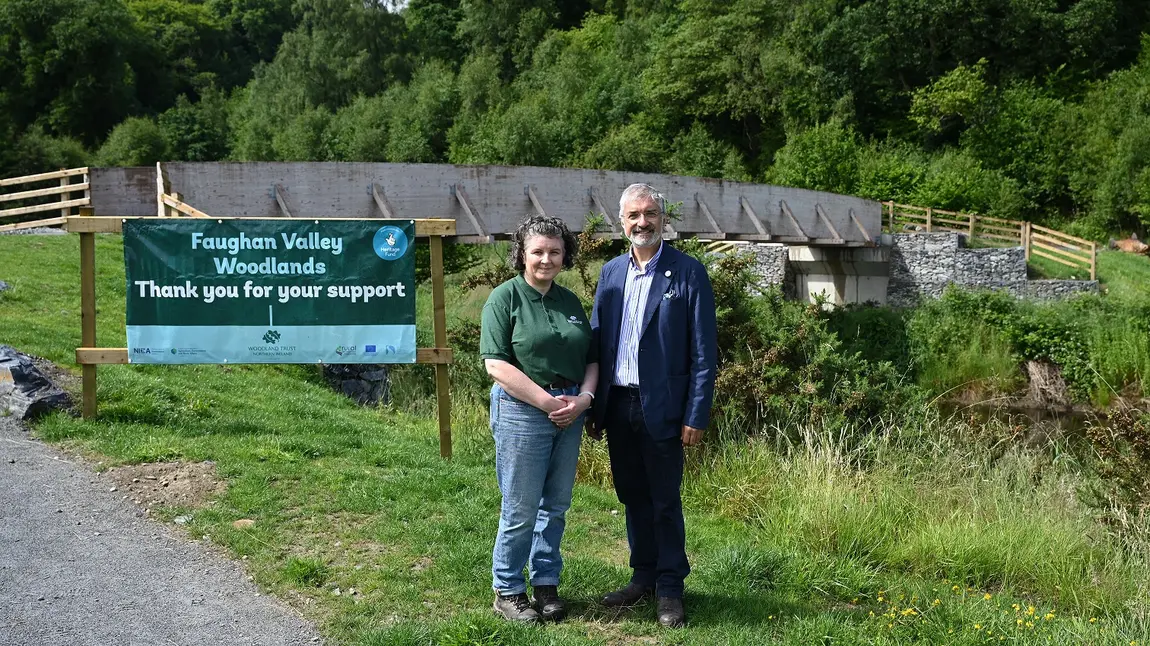 Two people are stood in front of the new bridge in Faughan Valley woodlands. Next to them is a sign to introduce the woodlands and has the Heritage Fund logo on it.