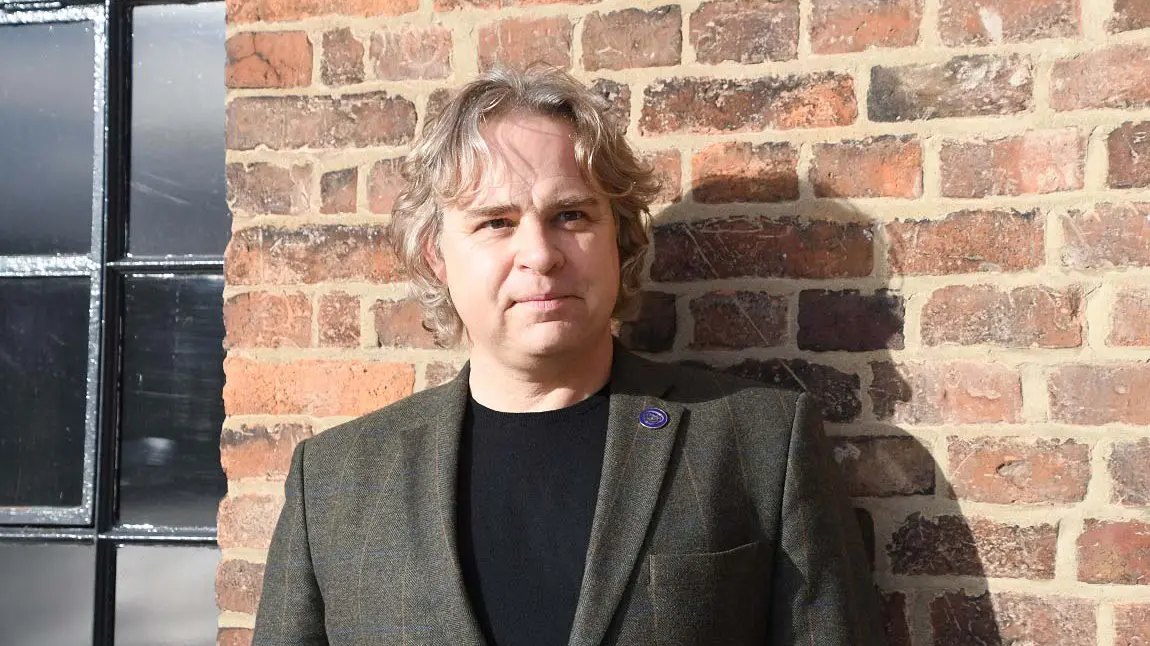 A person wearing a black and grey suit, standing in front of a red brick wall