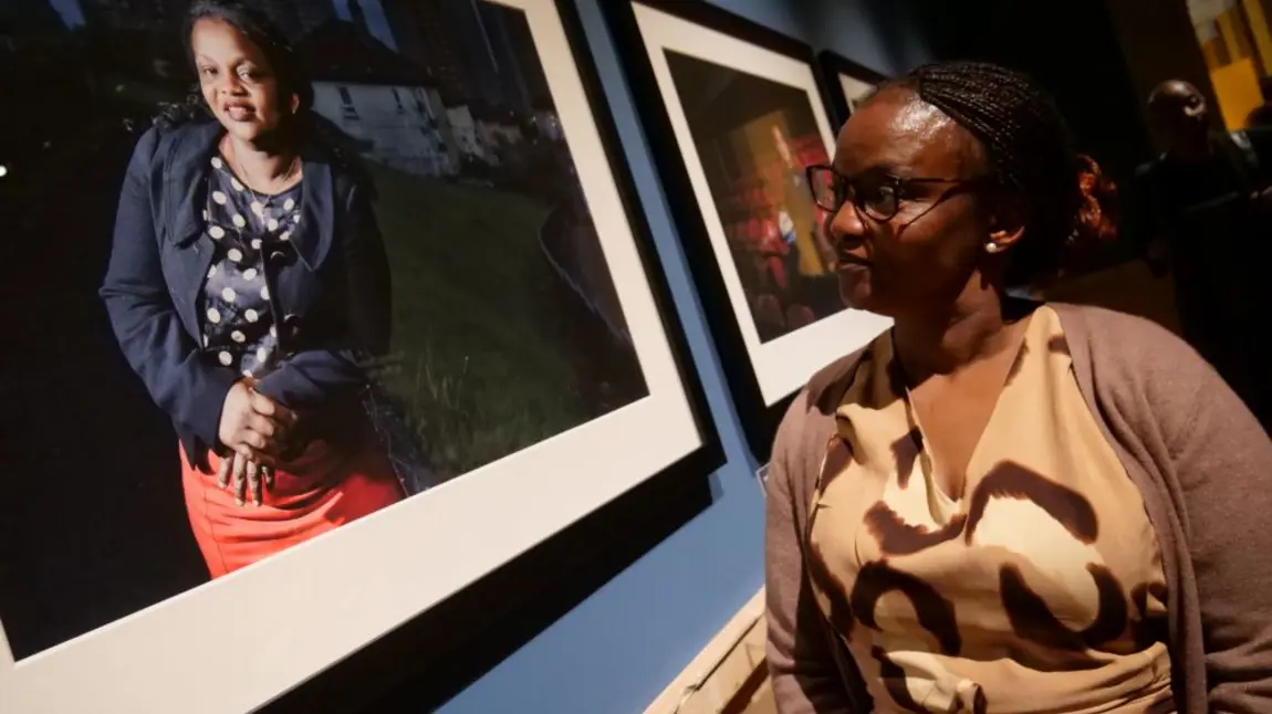 On the left is a framed photograph of a woman on the right there is a woman in a light brown dress looking at the picture