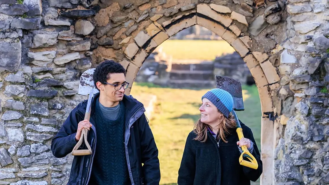 Two people holding shovels and dressed for cold weather walk through a doorway in a wall