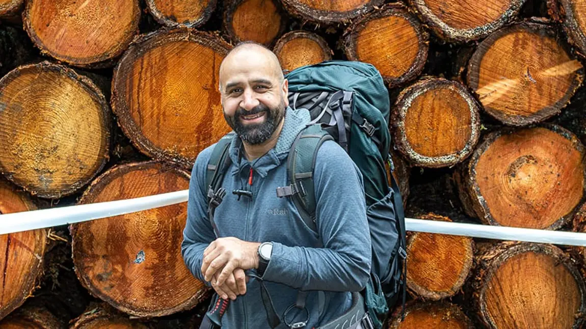 A man from a Pakistani family background on a outdoor walk, wearing hiking gear. He stands in front of a pile of logs