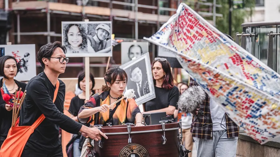Two people of Chinese heritage play a drum in a procession, while people in the background hold placards with photos of Chinese relatives