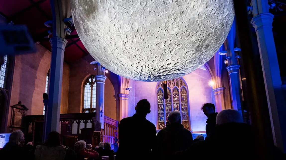 People look at a huge illuminated moon artwork suspended in a medium-size church