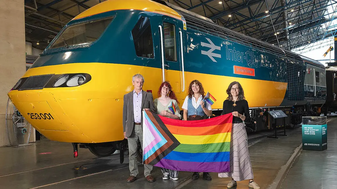 Four people stand in front of a train engine and hold a progress pride flag